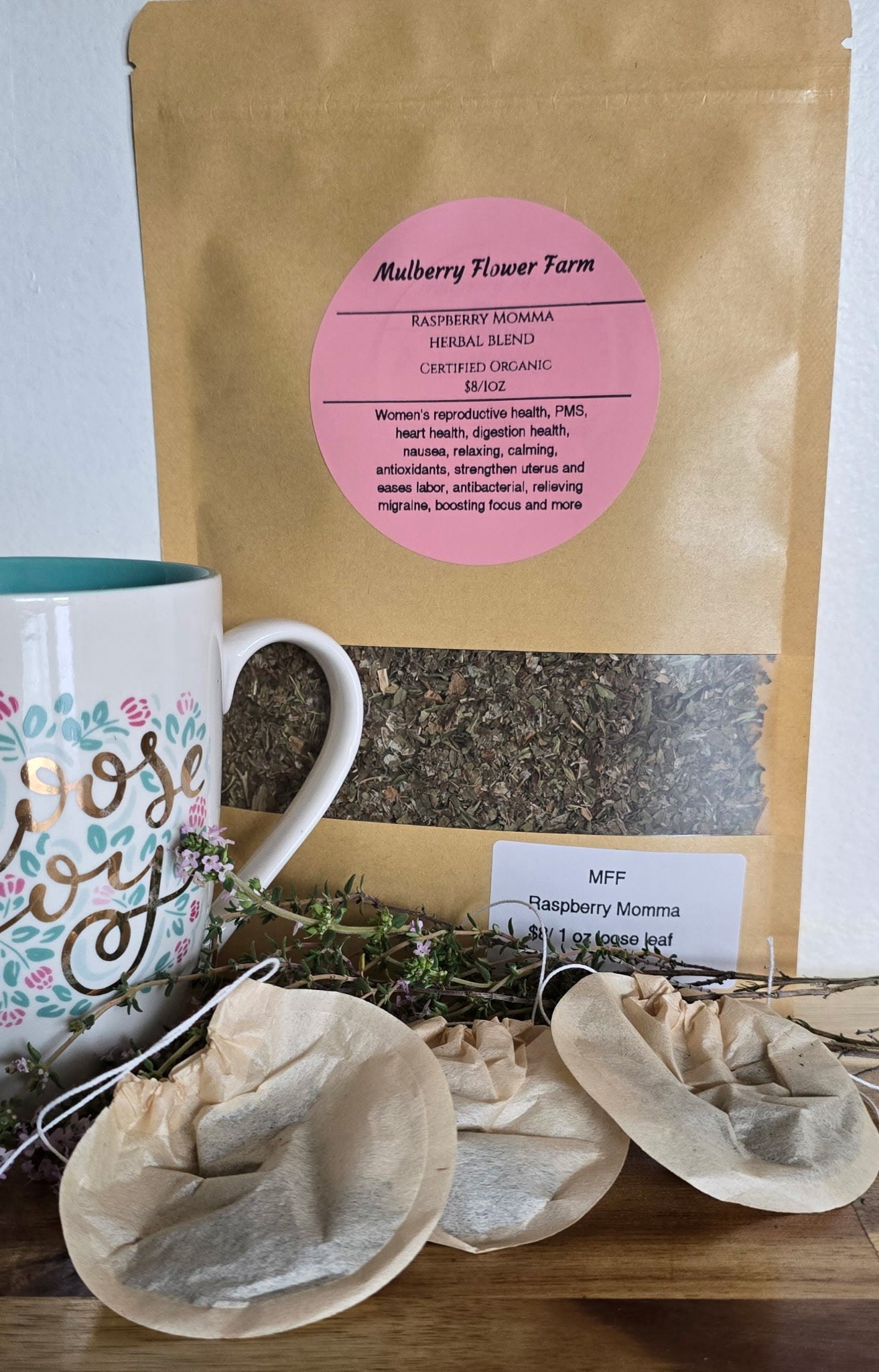 A brown paper bag from Mulberry Flower Farm has a pink circular label that reads "Raspberry Momma Herbal Blend" and "Certified Organic." The label mentions benefits such as women's reproductive health, PMS, heart health, digestion health, nausea, relaxing, calming, antioxidants, strengthening uterus, easing labor, antibacterial properties, relieving migraines, and boosting focus. A white mug with the words "choose joy" is partially visible. There are also some tea bags in the image.